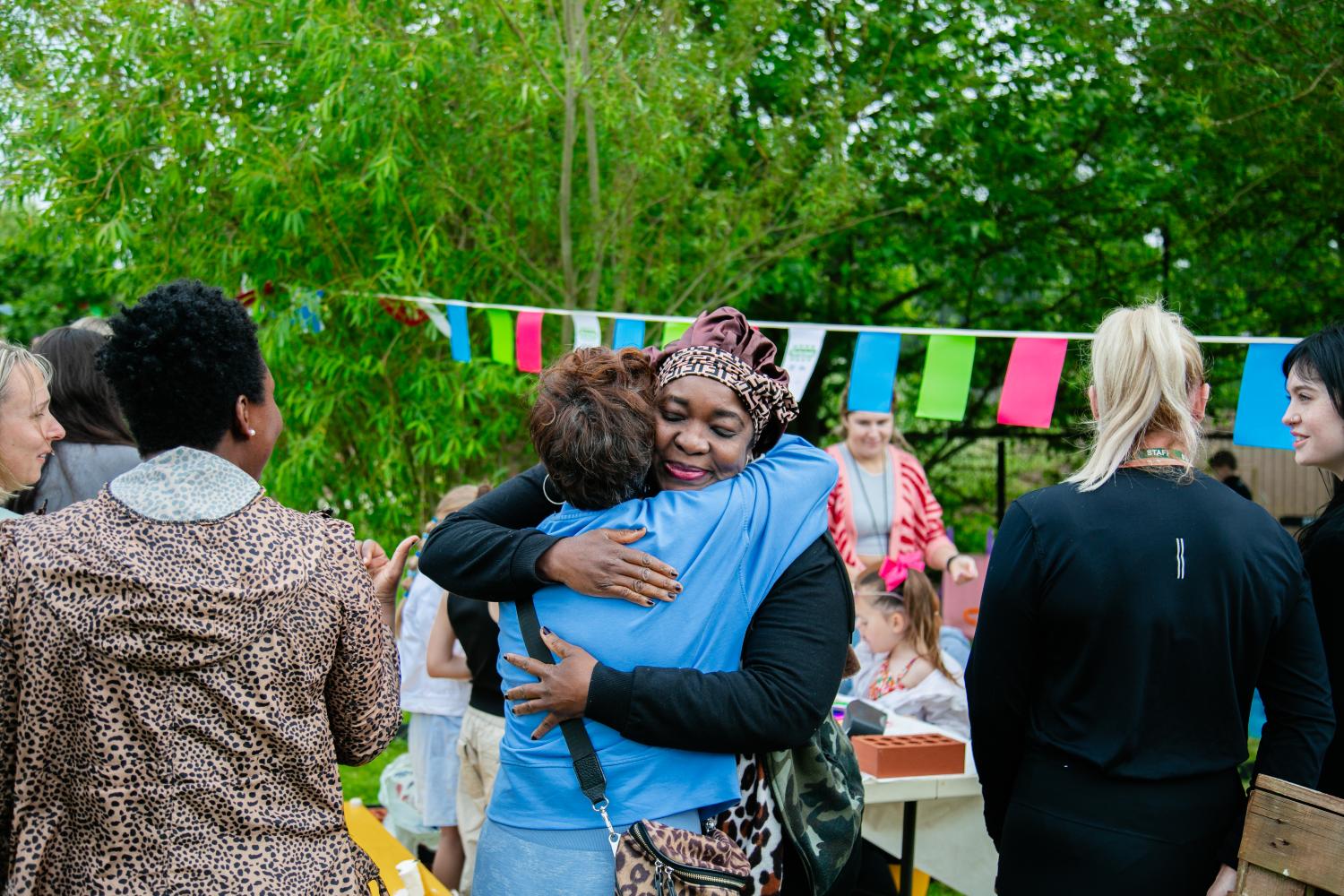 Two woman exchange a tender hug at an event. Lots of trees and bunting are visible in the background.