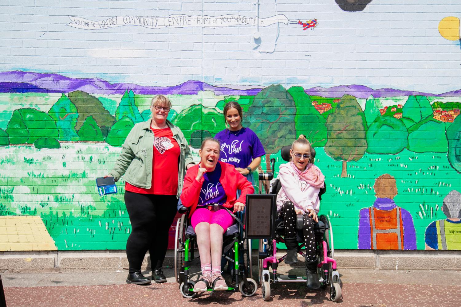 A group of women (two wheelchair users and two able bodied) stand in front of an incredible green mural smiling at the camera