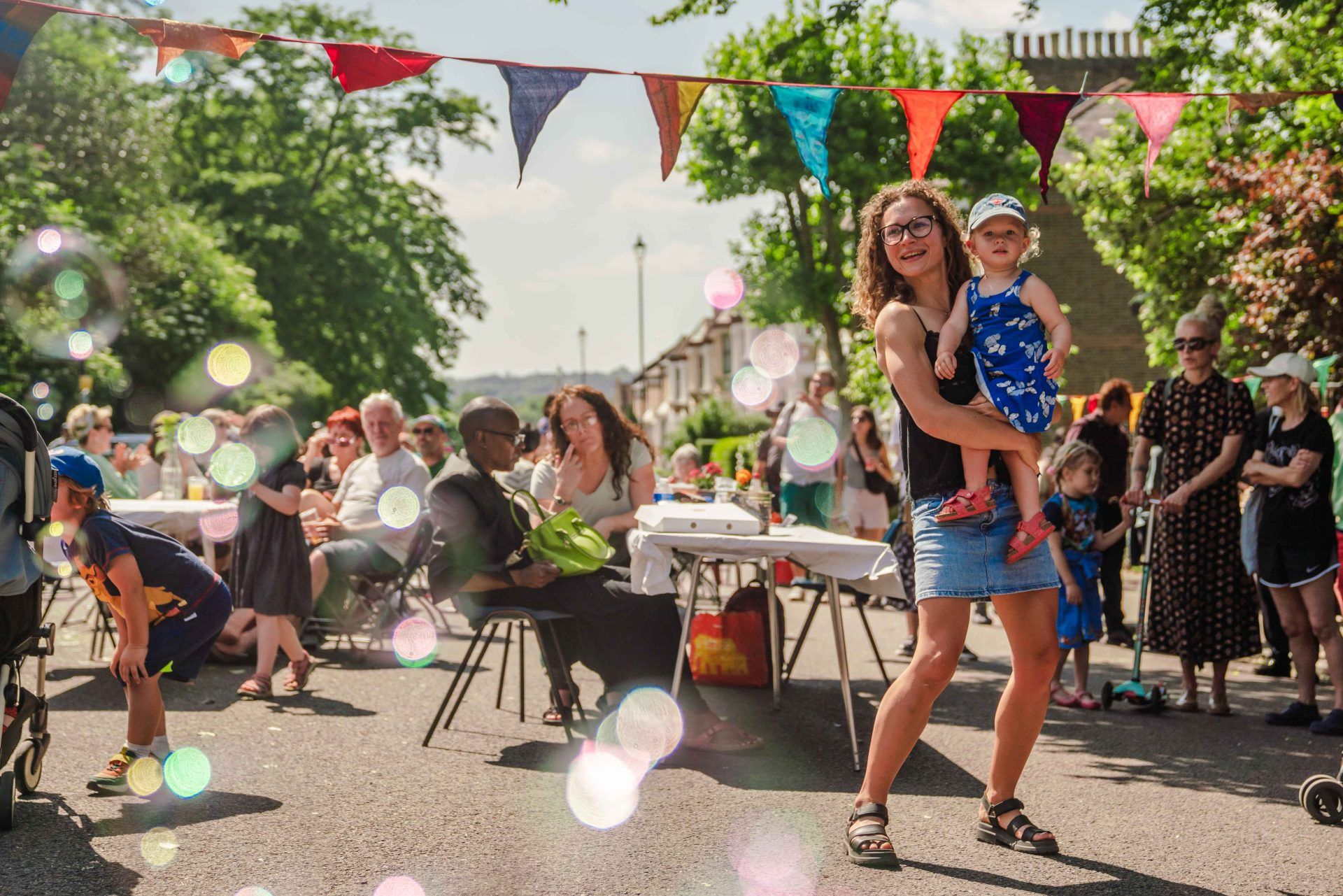 Woman holding a little girl at a street party. The sun is shining, tables are filled with chatting people and colourful bunting crosses the scene. New Cross Big Lunch 2024
