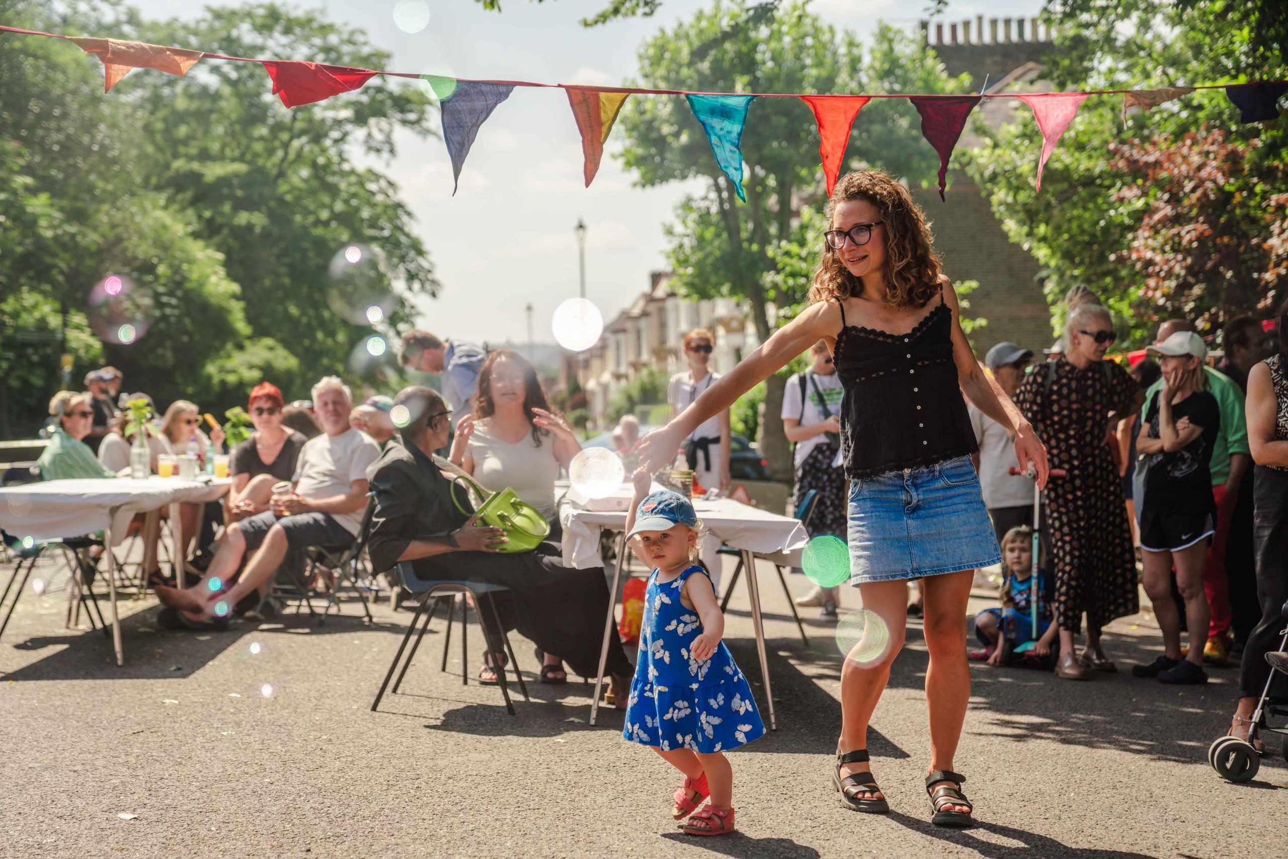 Woman dancing with a little girl at a street party. The sun is shining, tables are filled with chatting people and colourful bunting crosses the scene. New Cross Big Lunch 2024