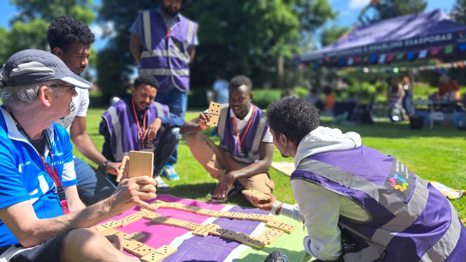 A group of men sitting wearing high-vis sitting and playing dominoes. They look like they're having a great time!