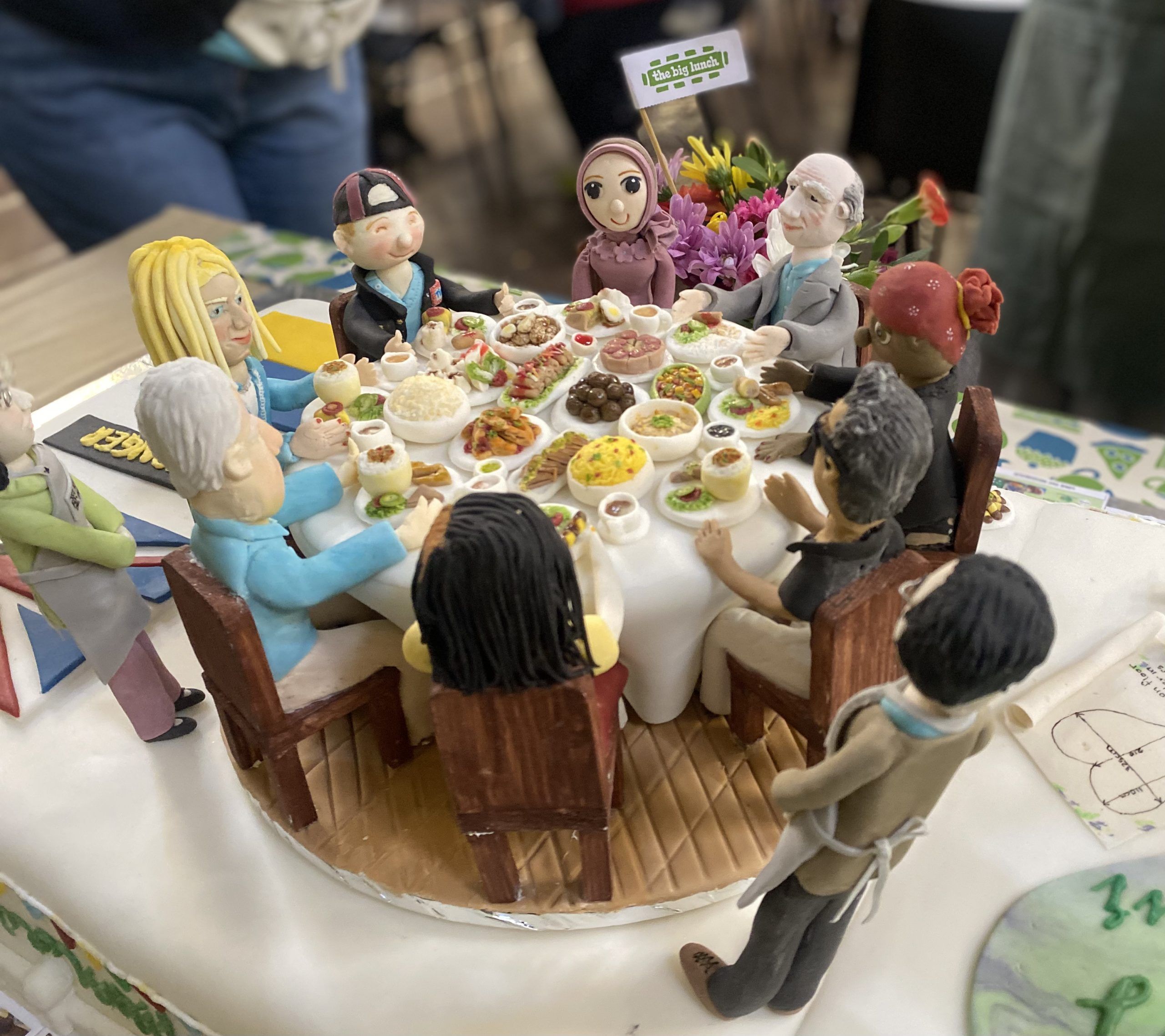An amazing cake with little figures sitting around a table laden with food and a Big Lunch flag