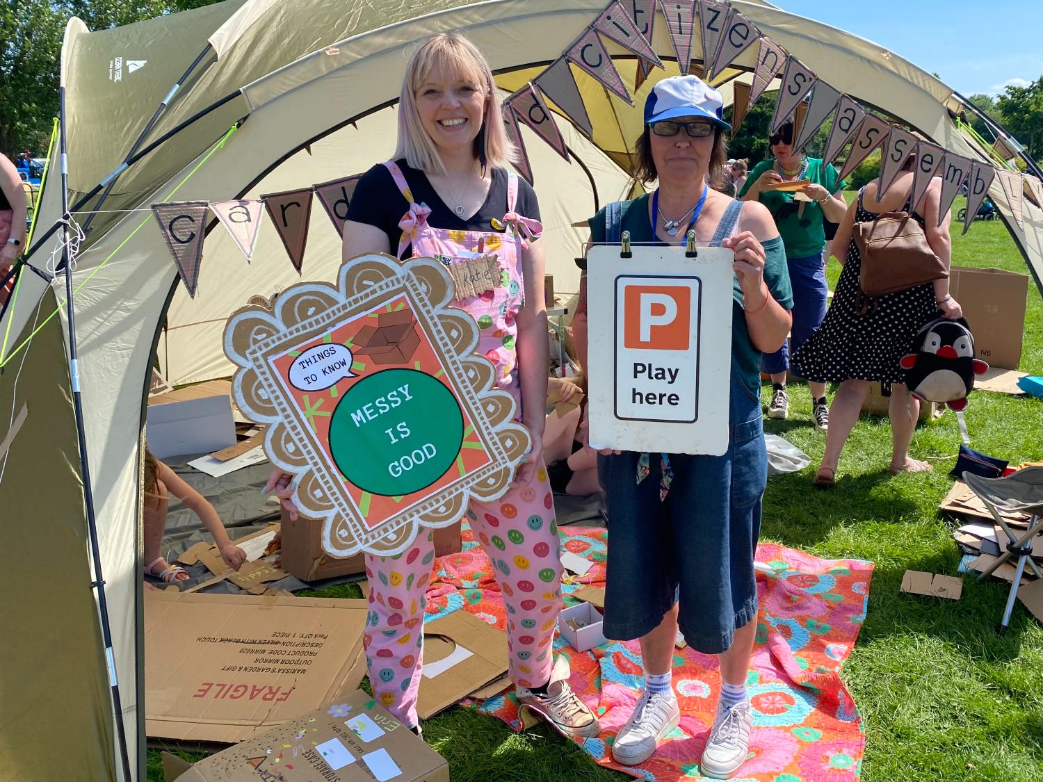 Two women hold up cardboard signs saying 'Messy is good' and 'Play here' with a gazebo full of crafts in the background
