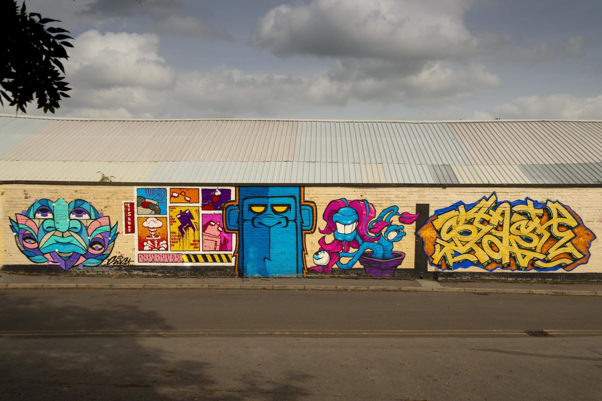 A brightly coloured wall of graffiti decorates a long brick wall next to GRIT Studios. The building behind has a corrugated roof in a clearly urban setting. The graffiti is skilfully done and displays an abstract face and what looks like an ape's face alongside some less discernible designs.